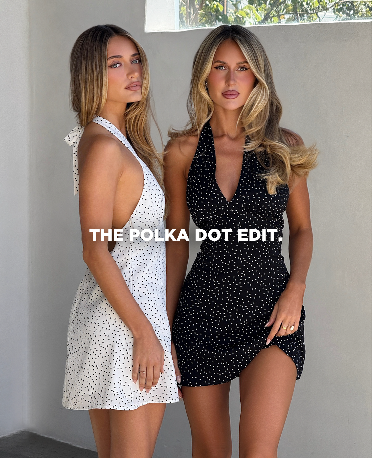 Two people standing in front of a white wall wearing halter dresses in opposite colours