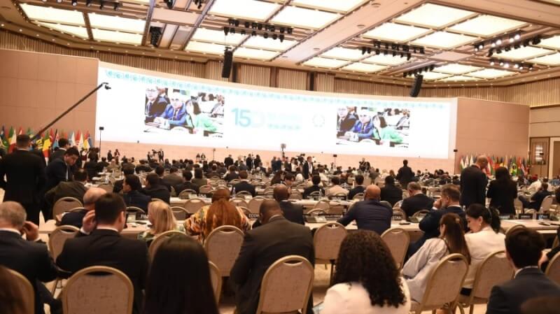 A wide-angle photo of the assembly from behind, showing participants and screens in the distance
