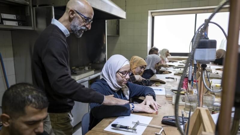 People sitting at desks in a row. The desks have curves cut out of them so they can sit sort of inside them, and they are working on something with their hands. A man is standing over them, helping them