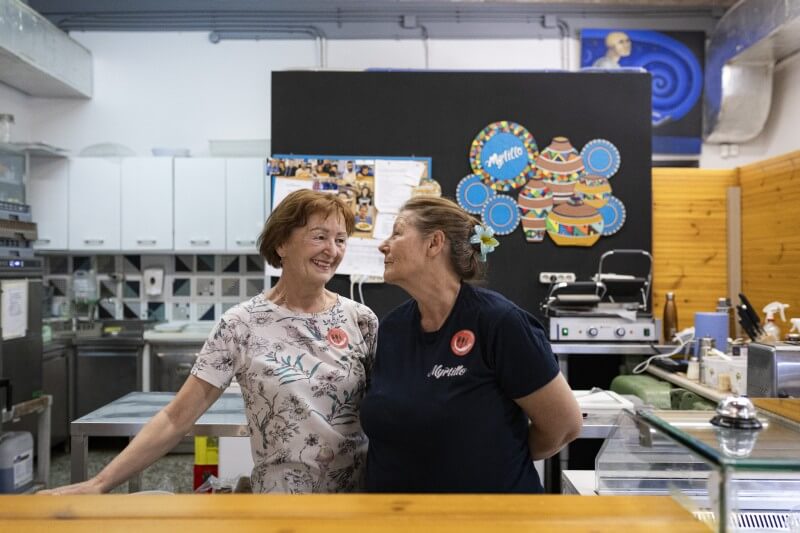 Two women in the kitchen of a restaurant, one is turning to the other and they both look happy
