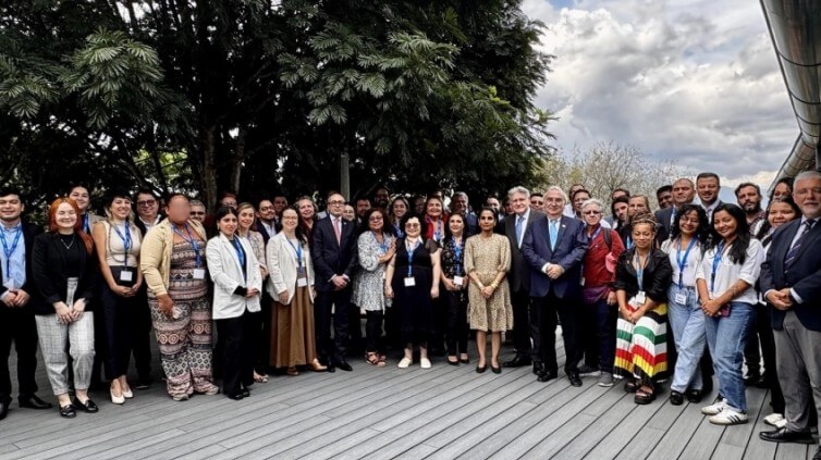 A group photo of around 50 people standing on some wooden decking. There is a tree directly behind those to the left, and behind those on the right is a cloudy, but bright sky.