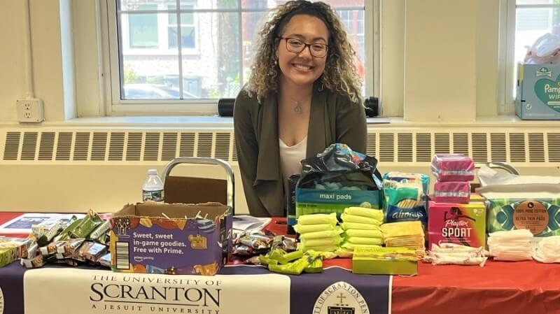 A woman sits at a booth with a Scranton logo. There are menstrual products and snacks on the table.