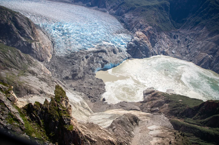 An aerial view of the end of a fjord, with the white-blue ice of a glacier at the top of the photo, surrounded by rocky walls, some of which have collapsed, leaving a swirl of muddy water