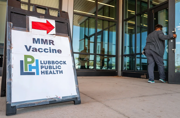 Sign for a Measles vaccination site outside an office building, as a woman opens the building door to go inside.