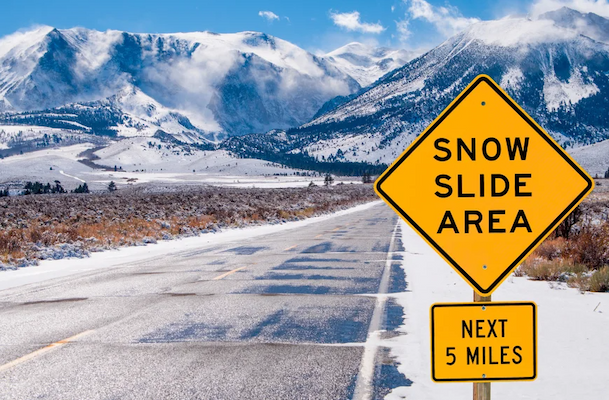 A yellow road sign reading "SNOW SLIDE AREA" next to a road, surrounded by snowy mountains.