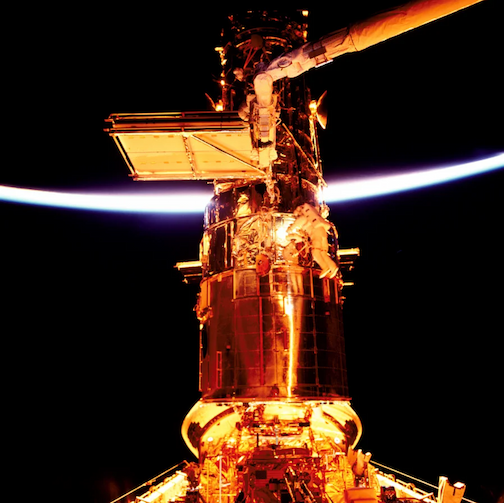 Two astronauts conducting a spacewalk alongside the exterior of the gold-tinted Hubble Space Telescope. The sunlit edge of Earth is visible in the background.