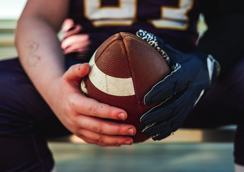 Close up of a young person a football, with one beat-up looking hand and one gloved hand.