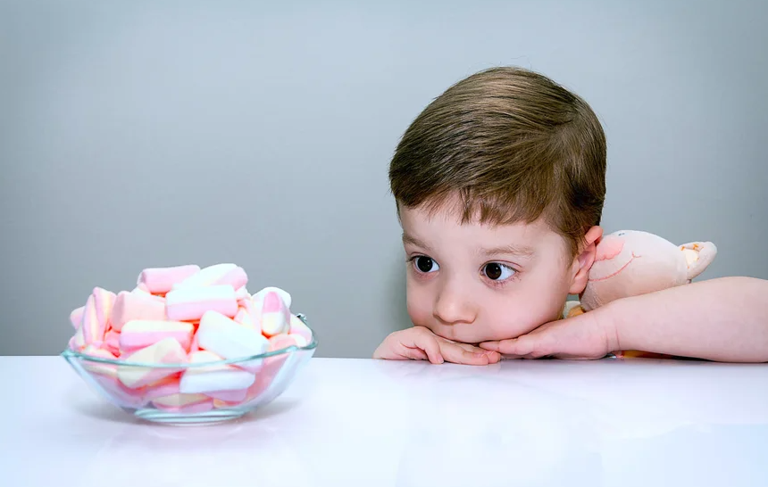 A little boy rests on the edge of a table staring at a bowl of pink and white candies