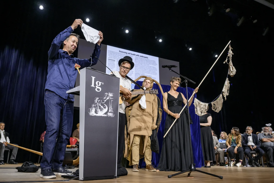 Researchers and performers onstage at the Ig Nobel Awards, with one presenter holding up a pair of underwear while others display a clothesline of undergarments.