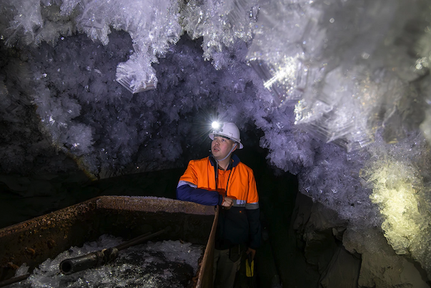 A man in a hardhat and headlamp stands below white crystal structures in an underground setting.