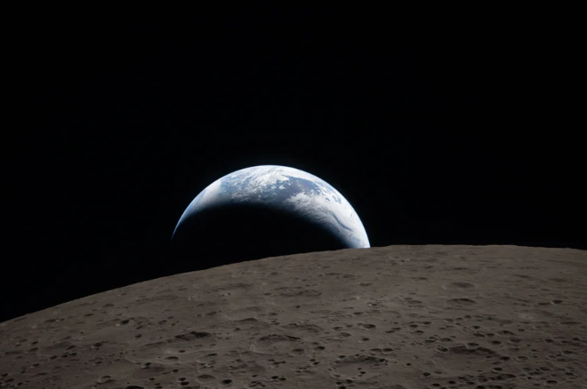 A crescent-lit Earth peeking out over a dark lunar surface like a half-obscured sunrise.