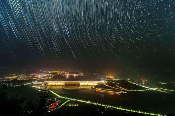 Long exposure photograph showing trails of stars over an illuminated dam and surrounding area