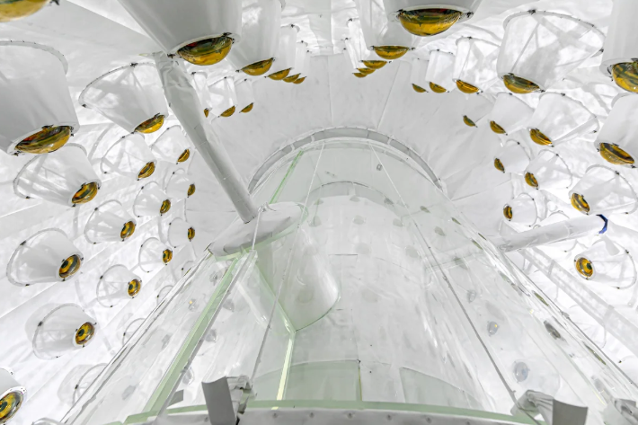 A giant cylindrical white vessel viewed at an angle from below, surrounded by a cylinder of cameras to collect light.