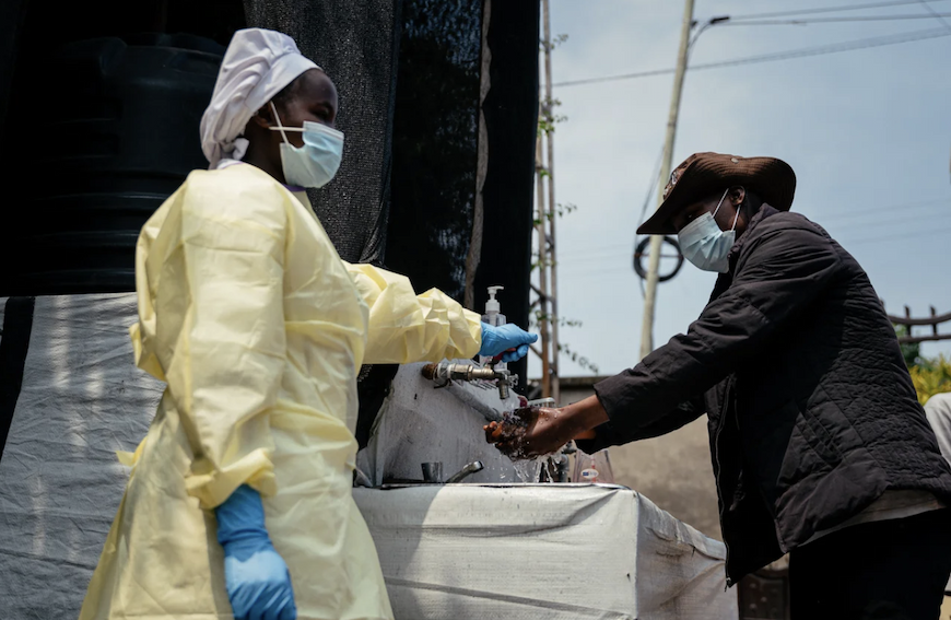 A man washes his hands at an outdoor sink, aided by a health care worker wearing a yellow gown, blue gloves, and mask and bonnet.