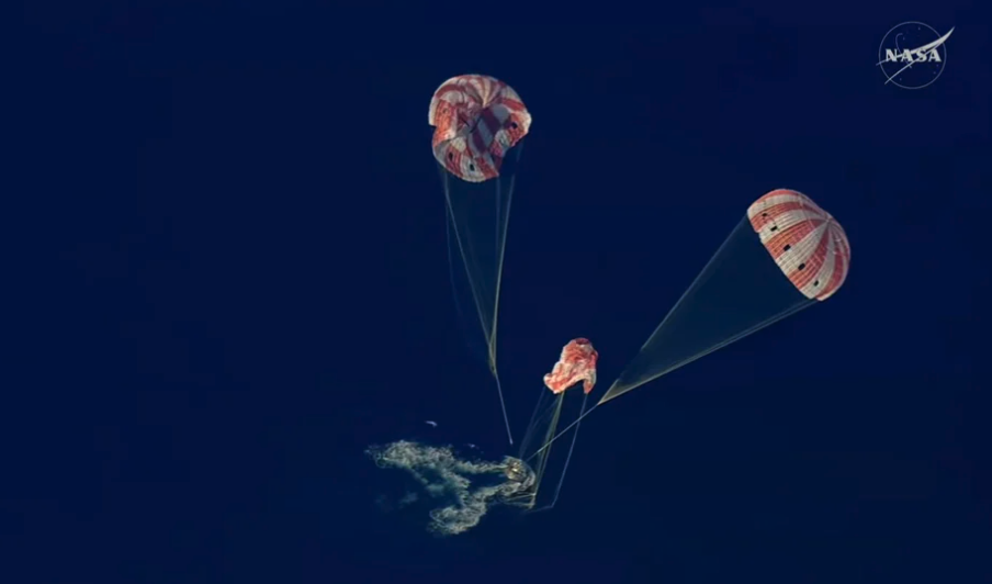 The Orion capsule splashes into the blue Pacific Ocean, with its three red-and-white parachutes in various states of collapse