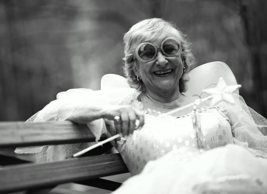 A gray-haired older lady sits on a park bench smiling. She wears a fairy costume and big round sunglasses and is holding a fairy wand.