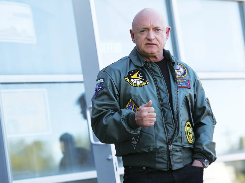Senator Mark Kelly in a green flight jacket giving a partial thumbs up