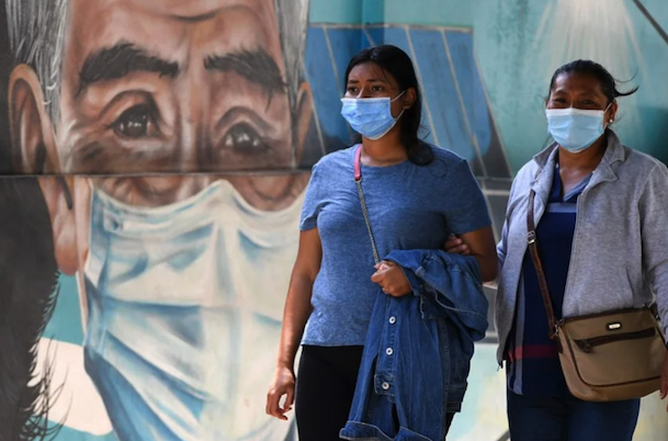Women wearing masks walk arm-in-arm in front of a mural of an elderly man wearing a mask.