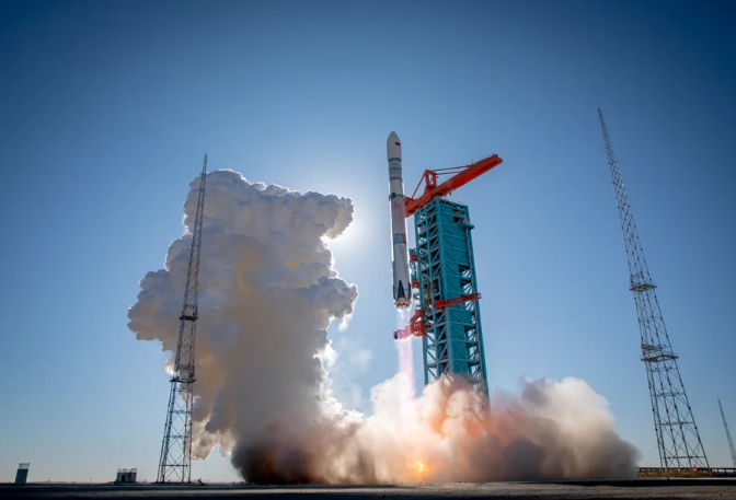 A photo of a rocket blasting off from a launch pad in a white-ish and fiery plume of exhaust under a clear, sunny sky.