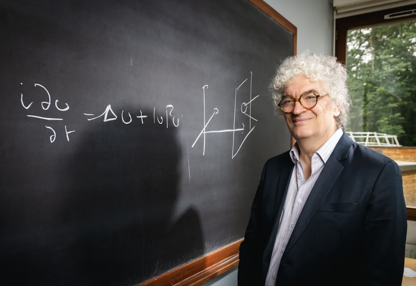 A man with curly white hair, wearing round spectacles, stands in front of equations on a blackboard.