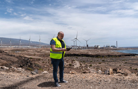 A man in a neon yellow vest looks at a tablet while standing on a brown landscape with several windturbines in the background.