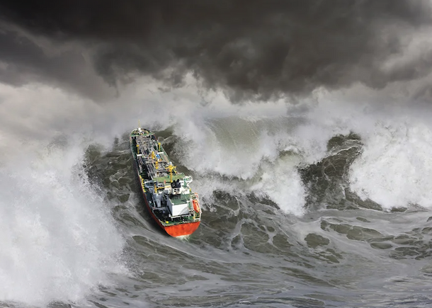 A cargo ship heading up the side of an aggressive wave in the ocean.
