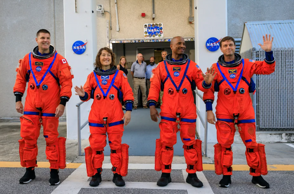The four astronauts of the Artemis II mission stand in a row in orange suits. Three are waving.