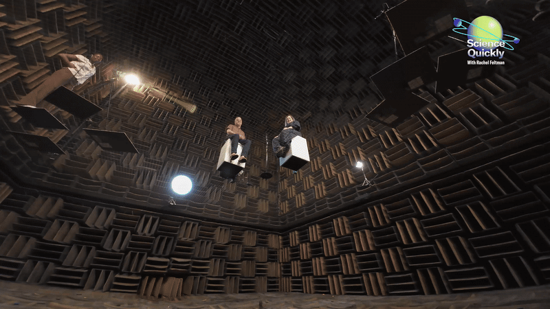A GIF of two people sitting on cubes that appear to float in the air inside a giant room lined with a brown grid.