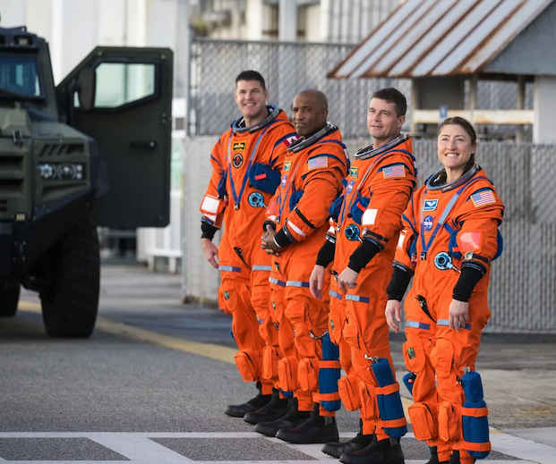 Four astronauts dressed in orange suits stand in a row, looking left and smiling at the camera