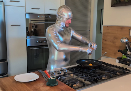 Person in a shiny silver full-body suit cooking at a stovetop, cracking eggs into a frying pan in a modern kitchen.