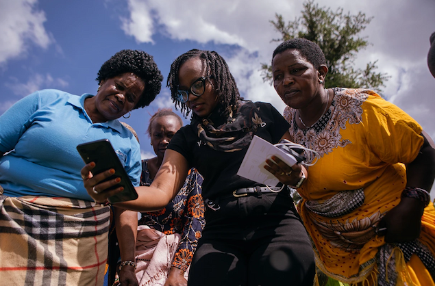 Four people in colorful outfits cluster together and all look down at a woman's phone, which she holds out at arm length under a blue sky.
