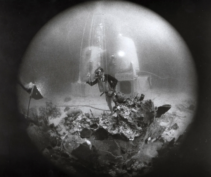 Black and white underwater photo of a diver on the seabed wearing a mask in front of a pod-like habitat