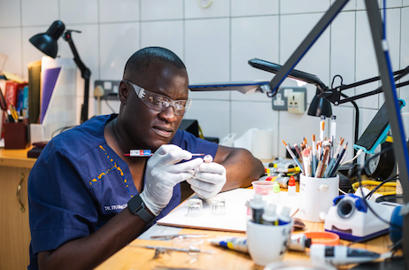 A researcher in dark blue scrubs and goggles works at a desk on something in his two gloved hands.