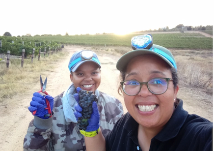 Two women in headlamps take a selfie while holding shears and a bunch of grapes next to a vineyard.