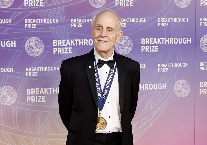 A man in a tuxedo wears a gold medal around his neck in front of a purple background that reads "Breakthrough Prize"