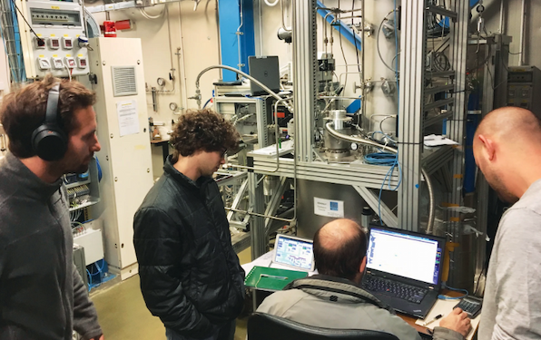 Researchers monitor instruments and computer screens in a laboratory filled with scientific equipment and cables.