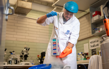 A man wearing a white lab coat, blue-capped face shield and gloves handles a tall box carrying multicolored cell samples