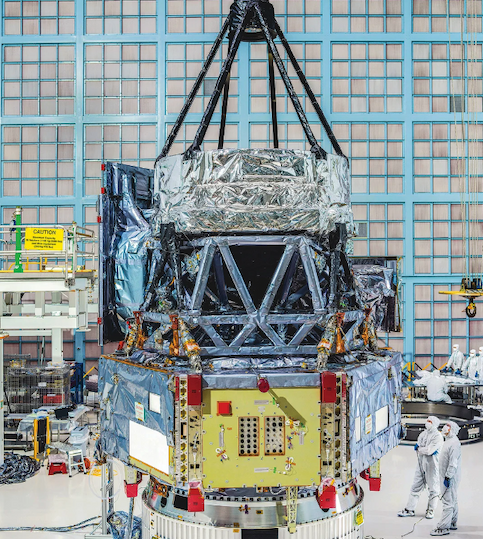 Large foil-wrapped space telescope components in a clean-room facility, with technicians in protective suits standing nearby.