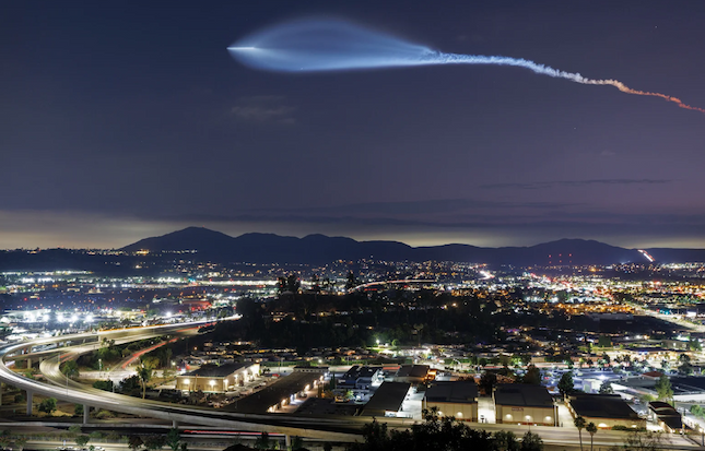 A rocket with white and red smoke streaks over an urban area at night.