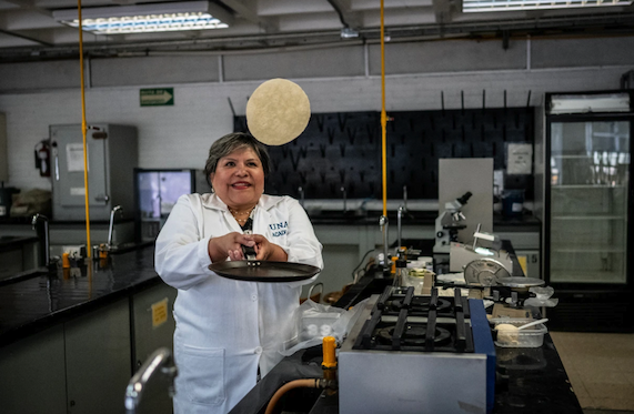Wearing a white lab coat while standing in a laboratory, Raquel Gómez Pliago holds a frying pan in two hands a flips a tortilla