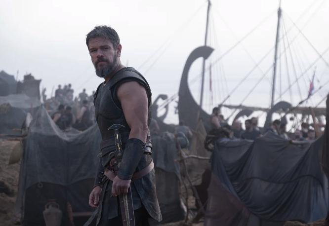 Promotional still from The Odyssey showing Odysseus, portrayed by Matt Damon, standing in armor with a sword in a busy coastal camp, with ships and soldiers visible behind him.