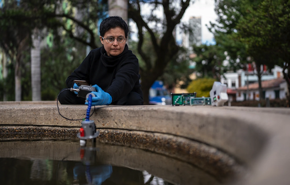 A person in a whilte protective suit, goggles, facemask and wearing blue gloves squats to take a sample in the rocky and snowy Antarctic landscape