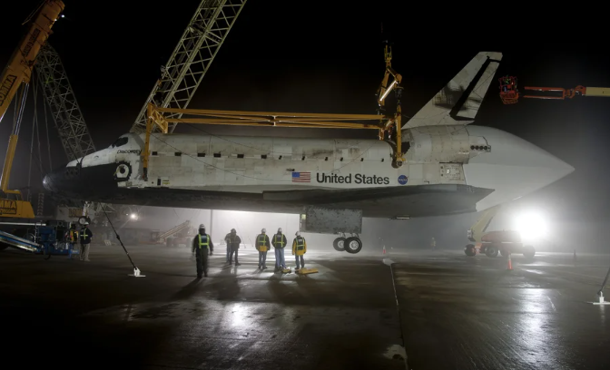 Photo of a space shuttle hoisted in air by a specialized crane holding it above a landing strip as several hard-hatted workers observe.