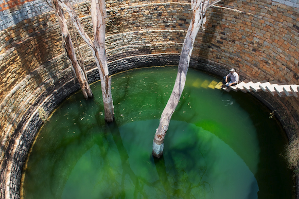 A man in a ballcap sits on floating stone stairs that descend into a round brick well filled partially with green water and three tree trunks.