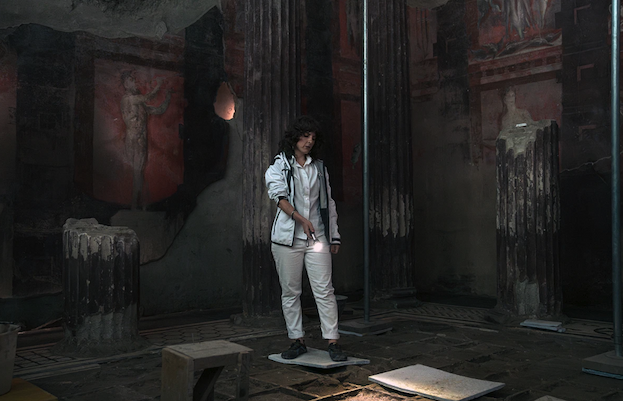 A woman in white pants and a white jacket points a flashlight at the ground inside ruins of Pompeii