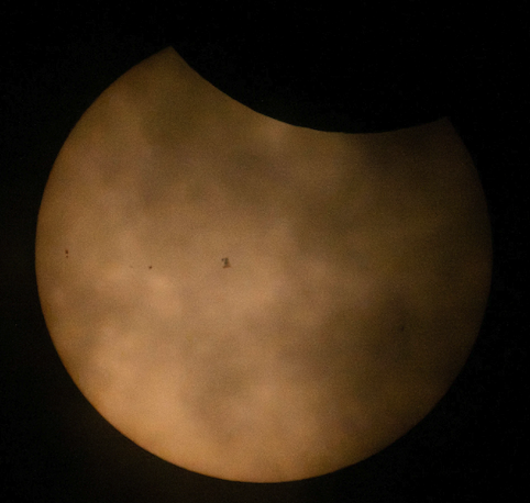 The sun during a partial solar eclipse, partially obscured by clouds, and the International Space Station is seen in silhouette at center as it transits across the sky