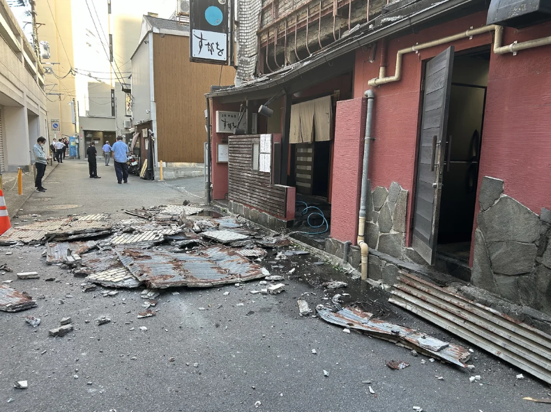 A photo looking down an alleyway where parts of a wall have collapsed onto the sidewalk. People stand around in the distance.
