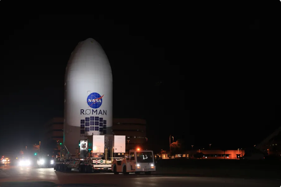 A large rocket fairing containing NASA's Nancy Grace Roman Space Telescope rests on a platform tugged by a truck, as it approaches integration with its rocket for launch.