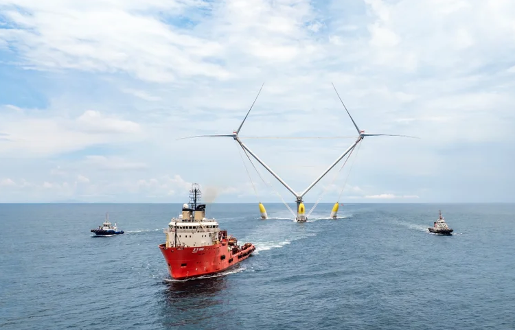 A red boat pulls an offshore wind turbine with two sets of blades anchored to a central platform over the ocean