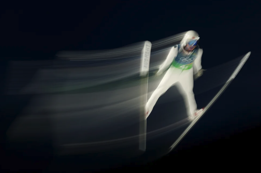A ski jumper in a white suit mid-jump behind a black sky.